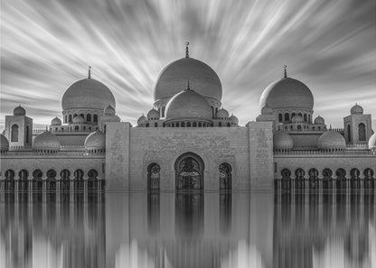 A stunning black and white long-exposure photograph of a grand mosque with multiple domes, reflected perfectly in calm water. The sky above is dramatic, with clouds streaking outwards, and the image is presented in a silver frame. Decor
