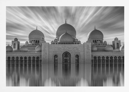 A stunning black and white long-exposure photograph of a grand mosque with multiple domes, reflected perfectly in calm water. The sky above is dramatic, with clouds streaking outwards, and the image is presented in a silver frame. Decor