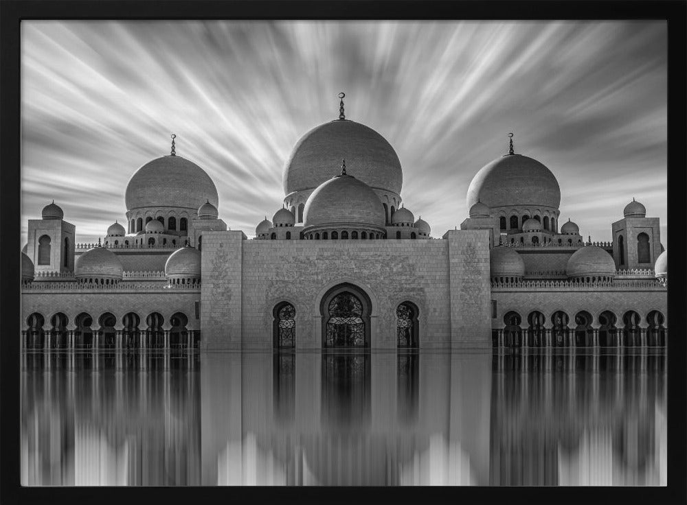 A stunning black and white long-exposure photograph of a grand mosque with multiple domes, reflected perfectly in calm water. The sky above is dramatic, with clouds streaking outwards, and the image is presented in a silver frame. Decor