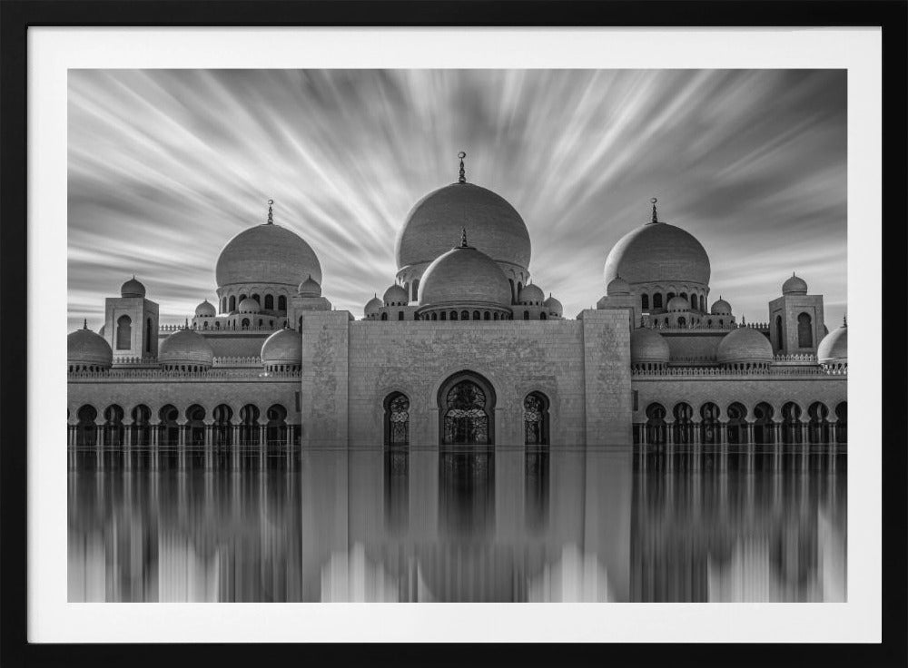 A stunning black and white long-exposure photograph of a grand mosque with multiple domes, reflected perfectly in calm water. The sky above is dramatic, with clouds streaking outwards, and the image is presented in a silver frame. Decor