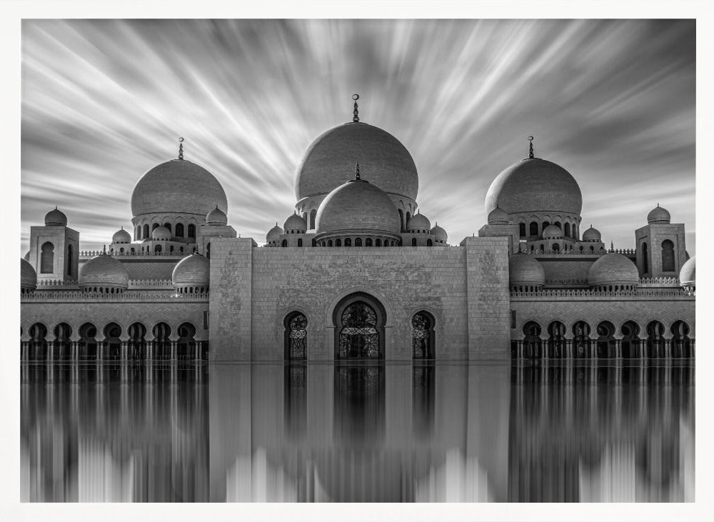 A stunning black and white long-exposure photograph of a grand mosque with multiple domes, reflected perfectly in calm water. The sky above is dramatic, with clouds streaking outwards, and the image is presented in a silver frame. Decor