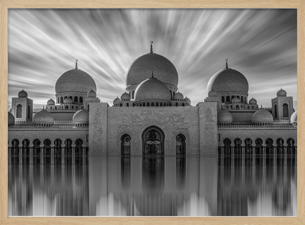A stunning black and white long-exposure photograph of a grand mosque with multiple domes, reflected perfectly in calm water. The sky above is dramatic, with clouds streaking outwards, and the image is presented in a silver frame. Decor