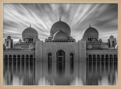 A stunning black and white long-exposure photograph of a grand mosque with multiple domes, reflected perfectly in calm water. The sky above is dramatic, with clouds streaking outwards, and the image is presented in a silver frame. Decor