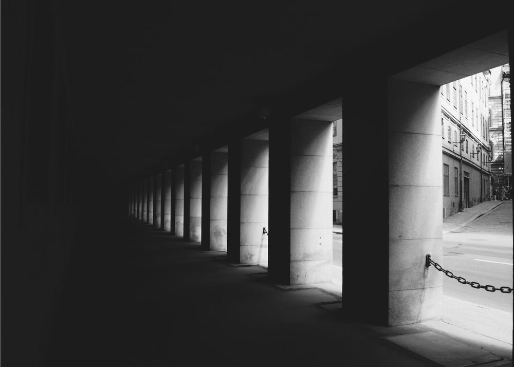 A high-contrast, black and white photograph from the perspective of a dark, covered walkway lined with a row of thick pillars. Light from an adjacent street highlights the pillars, creating a dramatic play of light and shadow and a strong sense of depth. Print