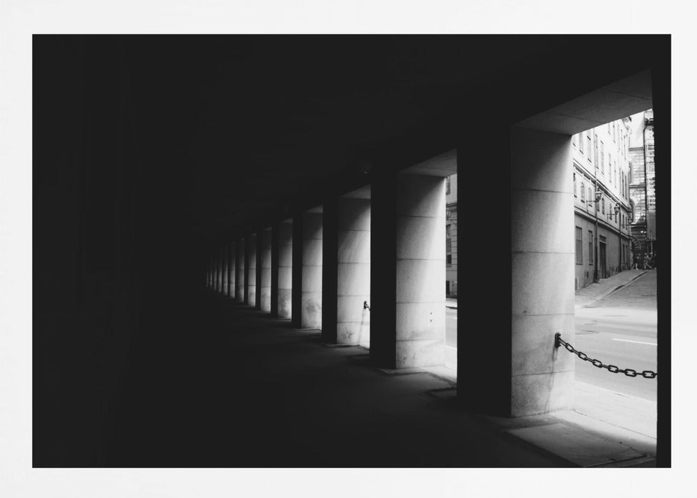 A high-contrast, black and white photograph from the perspective of a dark, covered walkway lined with a row of thick pillars. Light from an adjacent street highlights the pillars, creating a dramatic play of light and shadow and a strong sense of depth. Print
