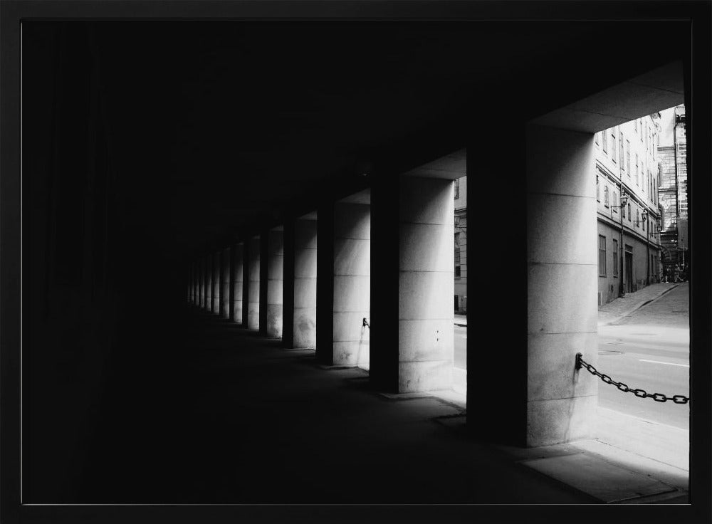 A high-contrast, black and white photograph from the perspective of a dark, covered walkway lined with a row of thick pillars. Light from an adjacent street highlights the pillars, creating a dramatic play of light and shadow and a strong sense of depth. Print