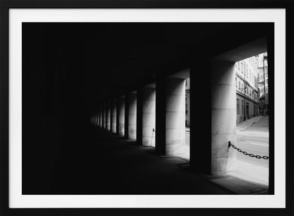 A high-contrast, black and white photograph from the perspective of a dark, covered walkway lined with a row of thick pillars. Light from an adjacent street highlights the pillars, creating a dramatic play of light and shadow and a strong sense of depth. Print