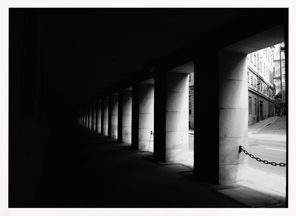A high-contrast, black and white photograph from the perspective of a dark, covered walkway lined with a row of thick pillars. Light from an adjacent street highlights the pillars, creating a dramatic play of light and shadow and a strong sense of depth. Print
