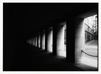 A high-contrast, black and white photograph from the perspective of a dark, covered walkway lined with a row of thick pillars. Light from an adjacent street highlights the pillars, creating a dramatic play of light and shadow and a strong sense of depth. Print