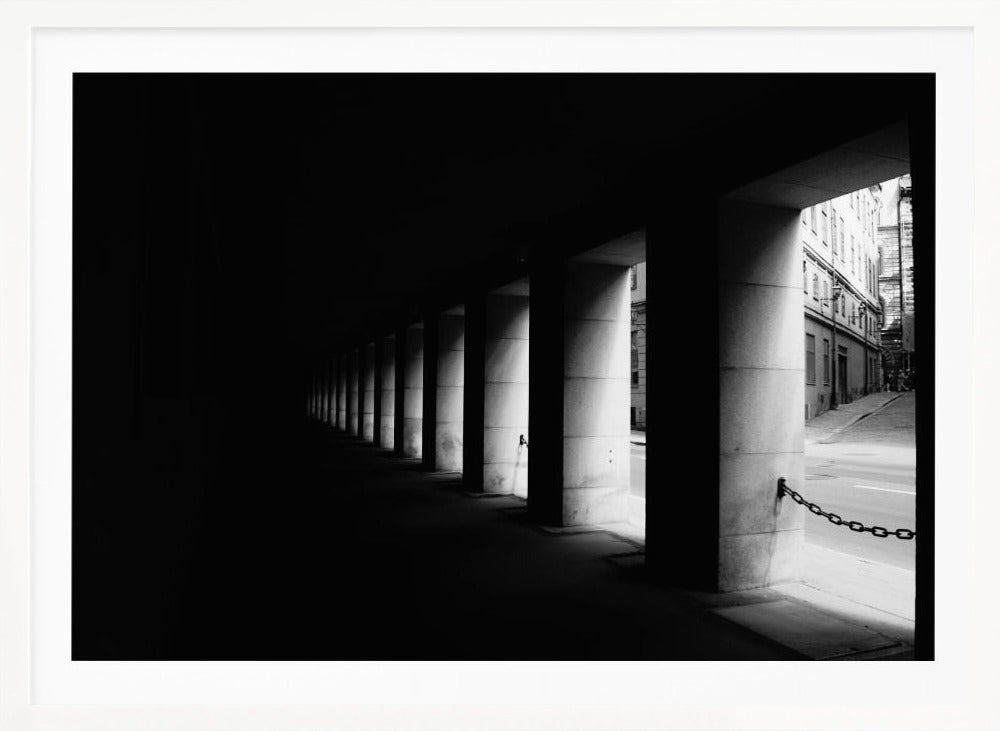 A high-contrast, black and white photograph from the perspective of a dark, covered walkway lined with a row of thick pillars. Light from an adjacent street highlights the pillars, creating a dramatic play of light and shadow and a strong sense of depth. Print