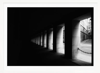 A high-contrast, black and white photograph from the perspective of a dark, covered walkway lined with a row of thick pillars. Light from an adjacent street highlights the pillars, creating a dramatic play of light and shadow and a strong sense of depth. Print