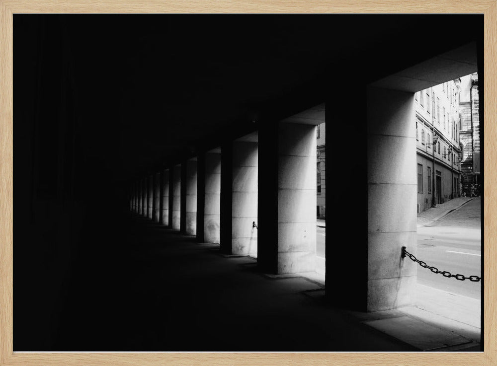 A high-contrast, black and white photograph from the perspective of a dark, covered walkway lined with a row of thick pillars. Light from an adjacent street highlights the pillars, creating a dramatic play of light and shadow and a strong sense of depth. Print