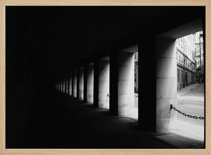 A high-contrast, black and white photograph from the perspective of a dark, covered walkway lined with a row of thick pillars. Light from an adjacent street highlights the pillars, creating a dramatic play of light and shadow and a strong sense of depth. Print