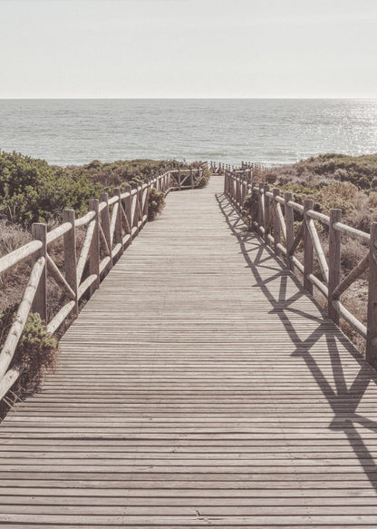 A long wooden boardwalk with rustic log railings leads the viewer's eye towards a calm, sunlit ocean under a hazy sky. Strong shadows from the railings fall across the path, creating a striped pattern. Poster
