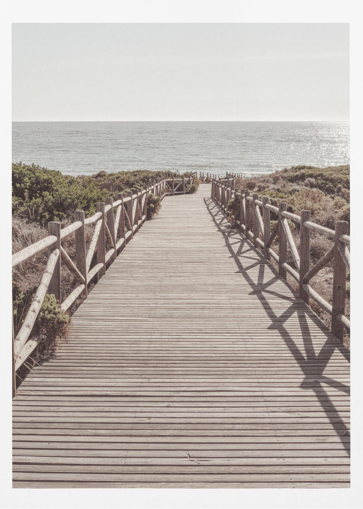A long wooden boardwalk with rustic log railings leads the viewer's eye towards a calm, sunlit ocean under a hazy sky. Strong shadows from the railings fall across the path, creating a striped pattern. Poster