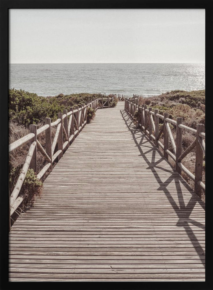 A long wooden boardwalk with rustic log railings leads the viewer's eye towards a calm, sunlit ocean under a hazy sky. Strong shadows from the railings fall across the path, creating a striped pattern. Poster