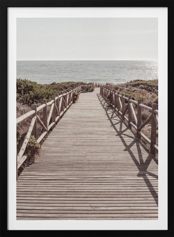 A long wooden boardwalk with rustic log railings leads the viewer's eye towards a calm, sunlit ocean under a hazy sky. Strong shadows from the railings fall across the path, creating a striped pattern. Poster