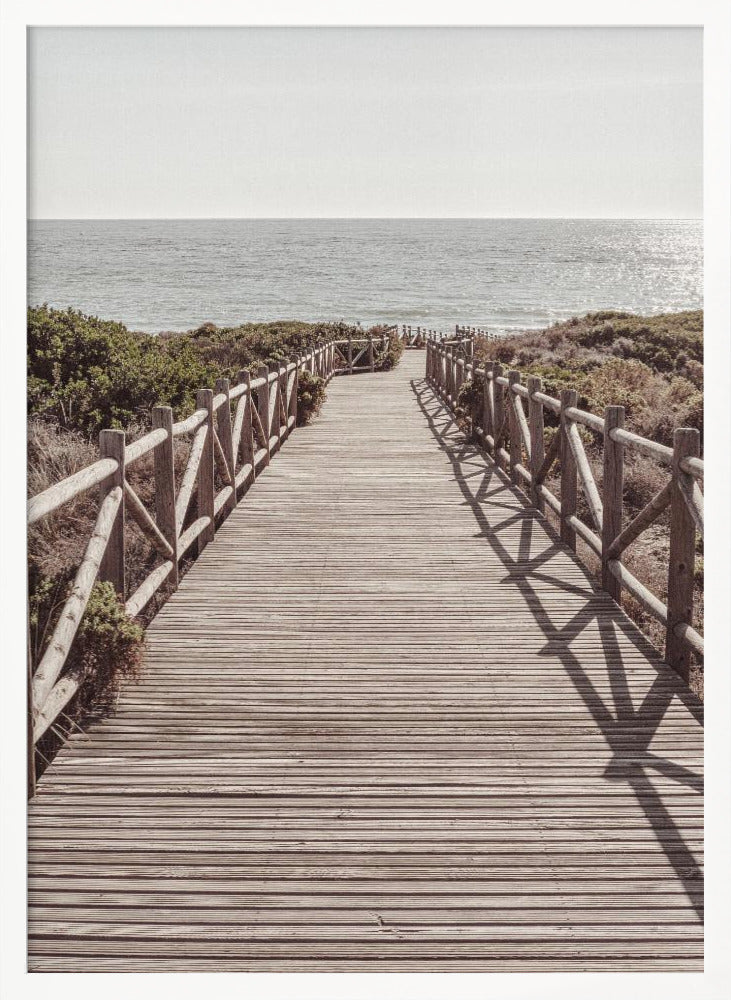 A long wooden boardwalk with rustic log railings leads the viewer's eye towards a calm, sunlit ocean under a hazy sky. Strong shadows from the railings fall across the path, creating a striped pattern. Poster