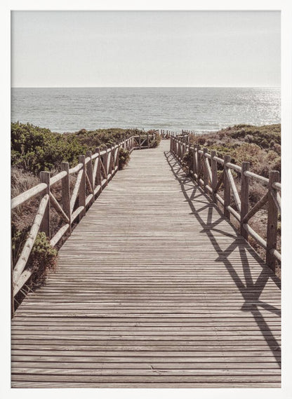 A long wooden boardwalk with rustic log railings leads the viewer's eye towards a calm, sunlit ocean under a hazy sky. Strong shadows from the railings fall across the path, creating a striped pattern. Poster