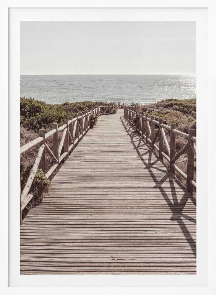 A long wooden boardwalk with rustic log railings leads the viewer's eye towards a calm, sunlit ocean under a hazy sky. Strong shadows from the railings fall across the path, creating a striped pattern. Poster