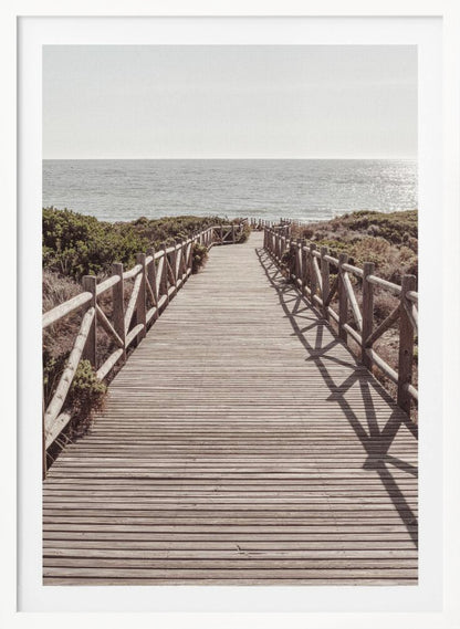 A long wooden boardwalk with rustic log railings leads the viewer's eye towards a calm, sunlit ocean under a hazy sky. Strong shadows from the railings fall across the path, creating a striped pattern. Poster