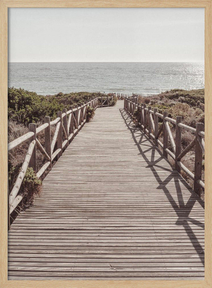 A long wooden boardwalk with rustic log railings leads the viewer's eye towards a calm, sunlit ocean under a hazy sky. Strong shadows from the railings fall across the path, creating a striped pattern. Poster