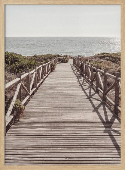 A long wooden boardwalk with rustic log railings leads the viewer's eye towards a calm, sunlit ocean under a hazy sky. Strong shadows from the railings fall across the path, creating a striped pattern. Poster