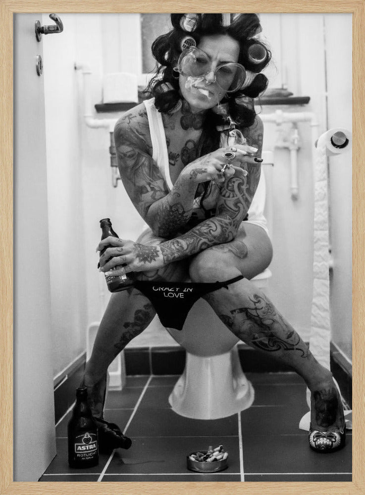 A black and white photo of a heavily tattooed woman with her hair in rollers sitting on a toilet. She wears large sunglasses, smokes a cigarette, holds a beer bottle, and has on black underwear that says 'CRAZY IN LOVE'. An ashtray and another beer bottle are on the floor. Artwork