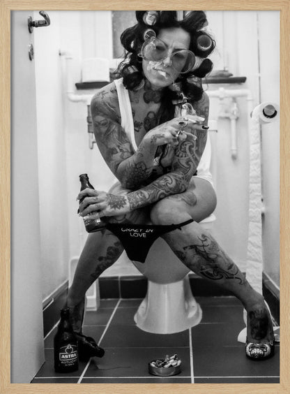 A black and white photo of a heavily tattooed woman with her hair in rollers sitting on a toilet. She wears large sunglasses, smokes a cigarette, holds a beer bottle, and has on black underwear that says 'CRAZY IN LOVE'. An ashtray and another beer bottle are on the floor. Artwork