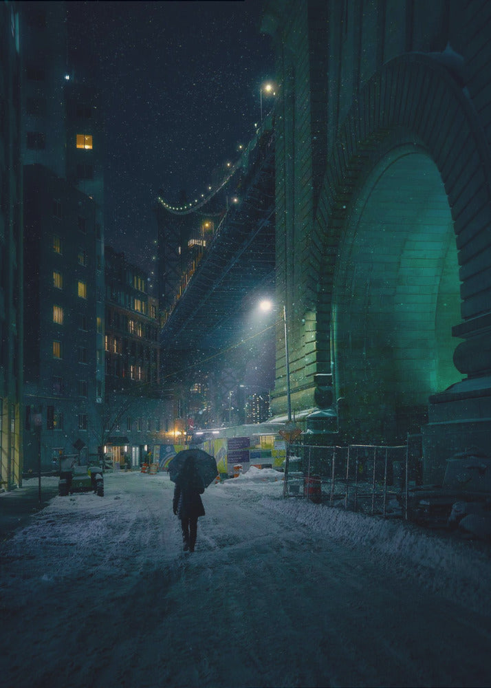 A lone person holding an umbrella walks down a snow-covered street at night, dwarfed by the massive, green-lit stone archway of the Brooklyn Bridge as snow falls around them. Print