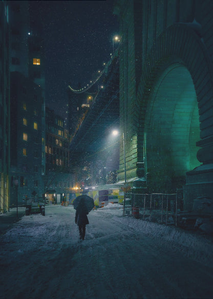 A lone person holding an umbrella walks down a snow-covered street at night, dwarfed by the massive, green-lit stone archway of the Brooklyn Bridge as snow falls around them. Print