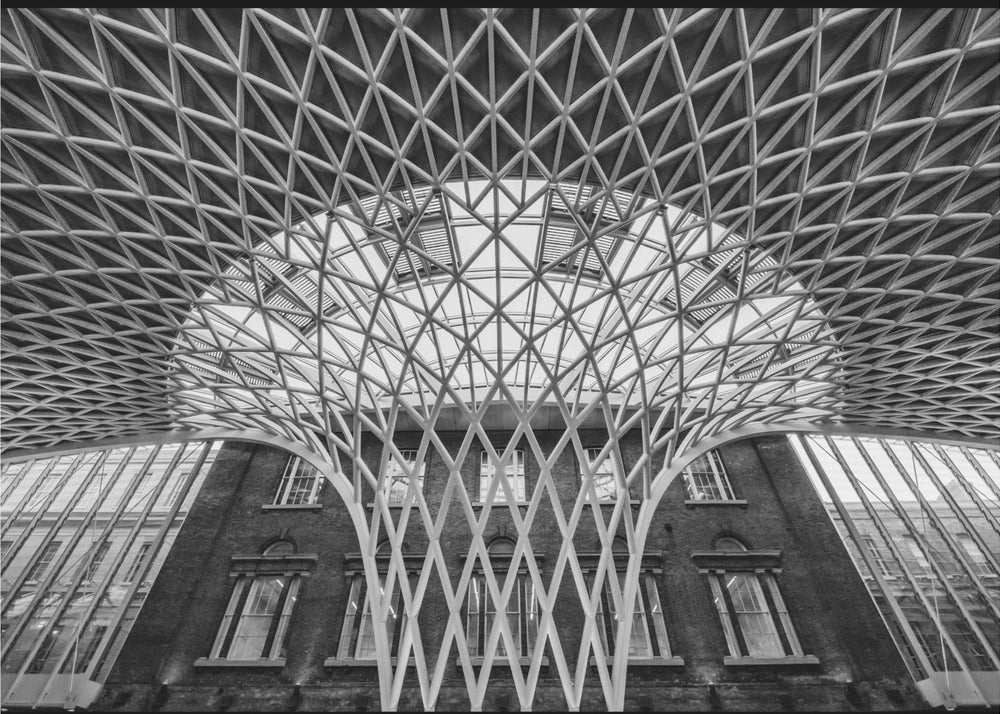 A framed, black and white photograph of the Western Concourse at King's Cross Station, London. The view is from a low angle, looking up at the intricate, white, funnel-like lattice roof structure as it spreads out across the ceiling, contrasting with the classic brick facade of the building behind it. Decor
