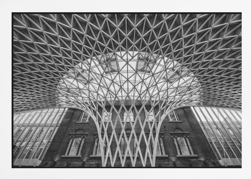 A framed, black and white photograph of the Western Concourse at King's Cross Station, London. The view is from a low angle, looking up at the intricate, white, funnel-like lattice roof structure as it spreads out across the ceiling, contrasting with the classic brick facade of the building behind it. Decor