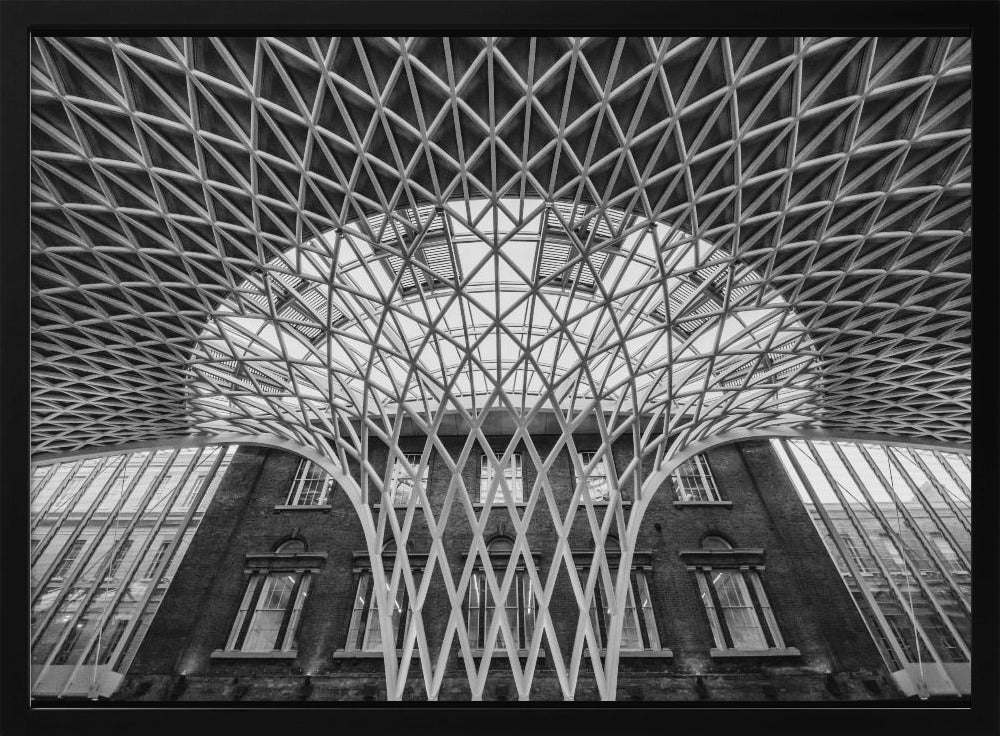 A framed, black and white photograph of the Western Concourse at King's Cross Station, London. The view is from a low angle, looking up at the intricate, white, funnel-like lattice roof structure as it spreads out across the ceiling, contrasting with the classic brick facade of the building behind it. Decor