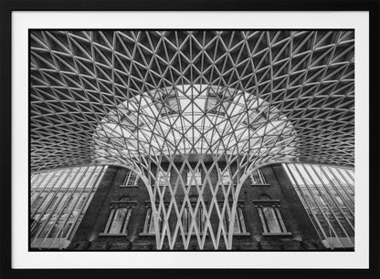 A framed, black and white photograph of the Western Concourse at King's Cross Station, London. The view is from a low angle, looking up at the intricate, white, funnel-like lattice roof structure as it spreads out across the ceiling, contrasting with the classic brick facade of the building behind it. Decor