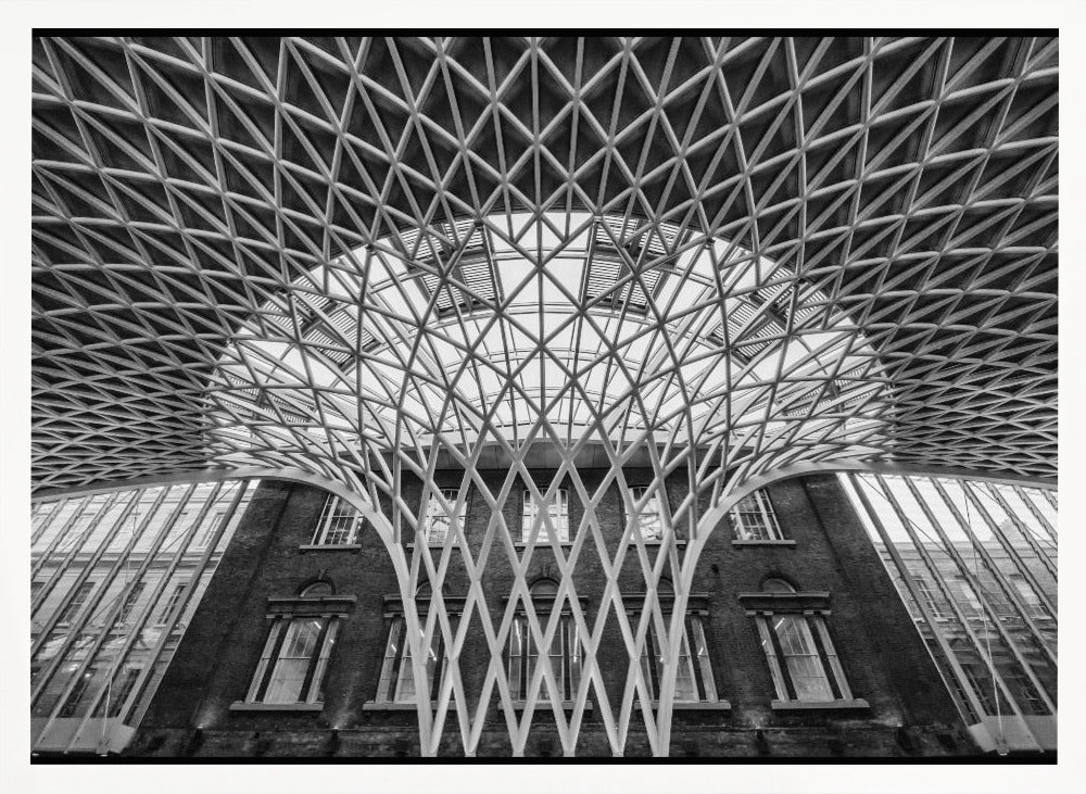 A framed, black and white photograph of the Western Concourse at King's Cross Station, London. The view is from a low angle, looking up at the intricate, white, funnel-like lattice roof structure as it spreads out across the ceiling, contrasting with the classic brick facade of the building behind it. Decor