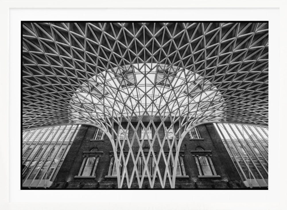 A framed, black and white photograph of the Western Concourse at King's Cross Station, London. The view is from a low angle, looking up at the intricate, white, funnel-like lattice roof structure as it spreads out across the ceiling, contrasting with the classic brick facade of the building behind it. Decor