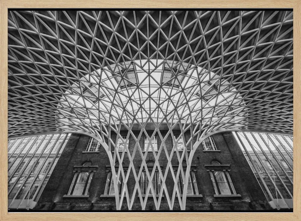 A framed, black and white photograph of the Western Concourse at King's Cross Station, London. The view is from a low angle, looking up at the intricate, white, funnel-like lattice roof structure as it spreads out across the ceiling, contrasting with the classic brick facade of the building behind it. Decor