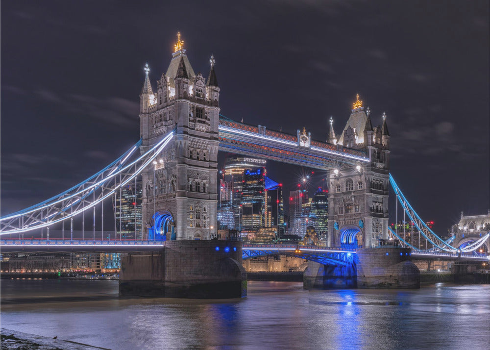 A vibrant night photograph of the iconic Tower Bridge in London, illuminated with bright white and electric blue lights. The dark River Thames in the foreground reflects the bridge and the lit-up modern city skyline in the background, all under a dark, cloudy sky. The image is enclosed in a silver frame. Decor