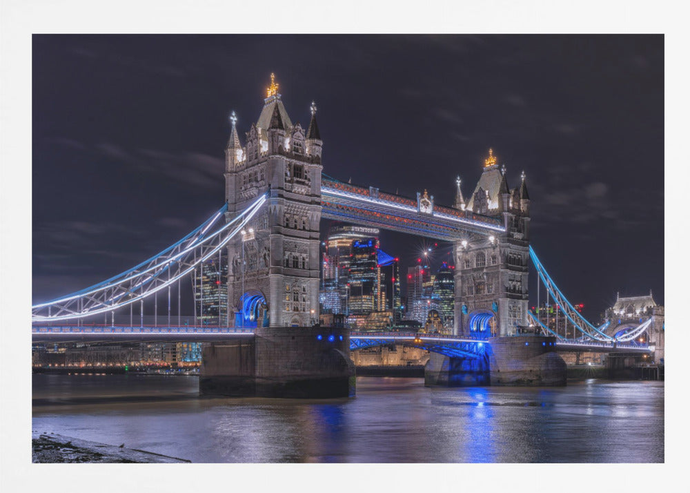 A vibrant night photograph of the iconic Tower Bridge in London, illuminated with bright white and electric blue lights. The dark River Thames in the foreground reflects the bridge and the lit-up modern city skyline in the background, all under a dark, cloudy sky. The image is enclosed in a silver frame. Decor