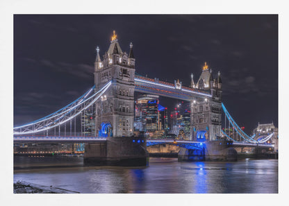 A vibrant night photograph of the iconic Tower Bridge in London, illuminated with bright white and electric blue lights. The dark River Thames in the foreground reflects the bridge and the lit-up modern city skyline in the background, all under a dark, cloudy sky. The image is enclosed in a silver frame. Decor