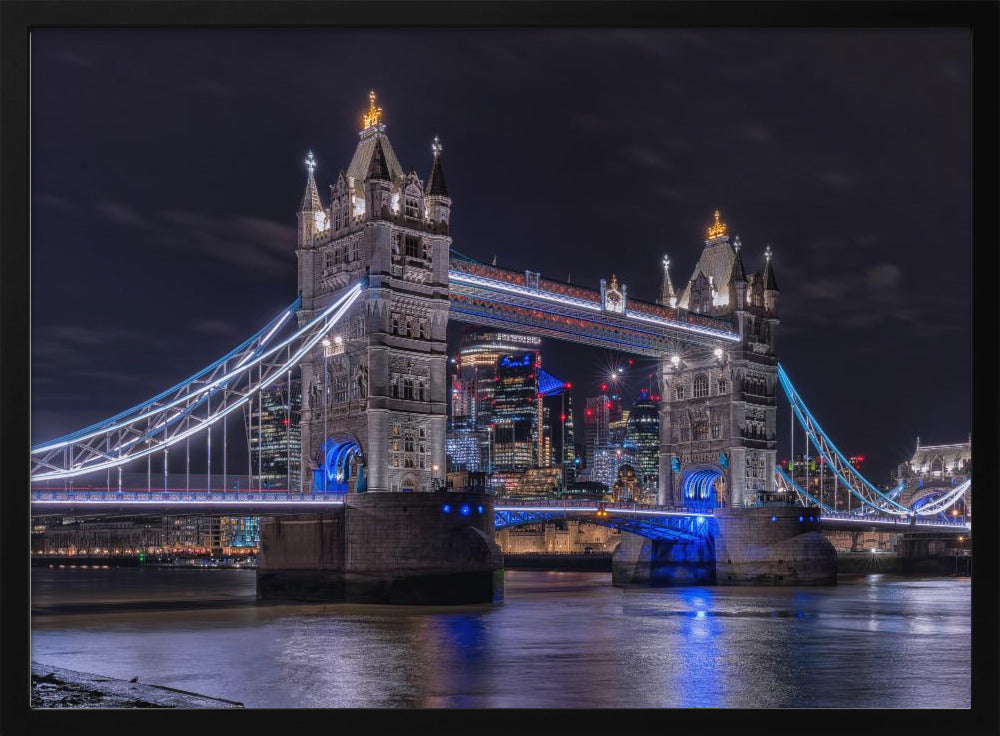 A vibrant night photograph of the iconic Tower Bridge in London, illuminated with bright white and electric blue lights. The dark River Thames in the foreground reflects the bridge and the lit-up modern city skyline in the background, all under a dark, cloudy sky. The image is enclosed in a silver frame. Decor