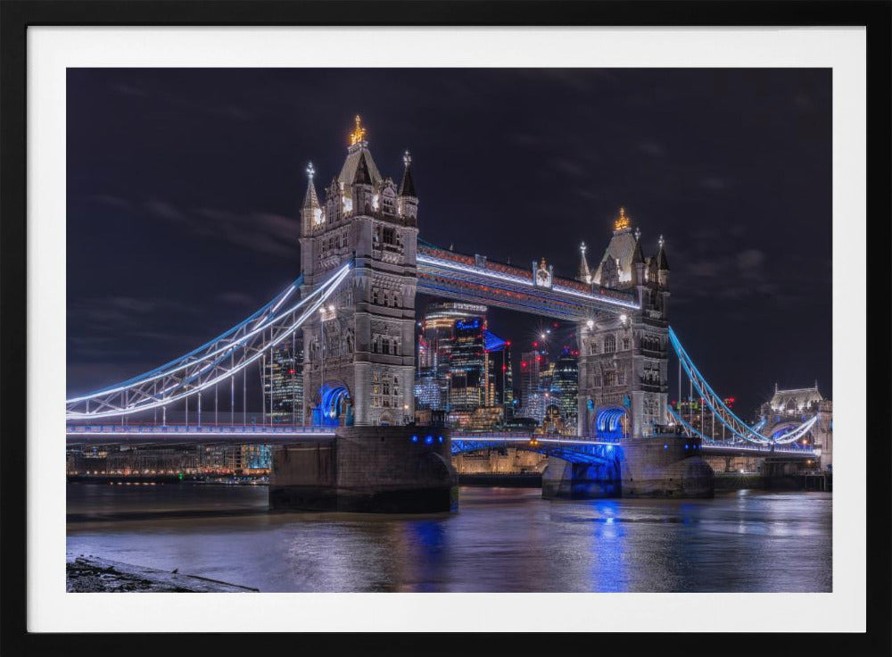 A vibrant night photograph of the iconic Tower Bridge in London, illuminated with bright white and electric blue lights. The dark River Thames in the foreground reflects the bridge and the lit-up modern city skyline in the background, all under a dark, cloudy sky. The image is enclosed in a silver frame. Decor