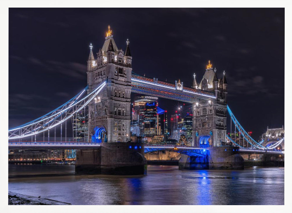 A vibrant night photograph of the iconic Tower Bridge in London, illuminated with bright white and electric blue lights. The dark River Thames in the foreground reflects the bridge and the lit-up modern city skyline in the background, all under a dark, cloudy sky. The image is enclosed in a silver frame. Decor
