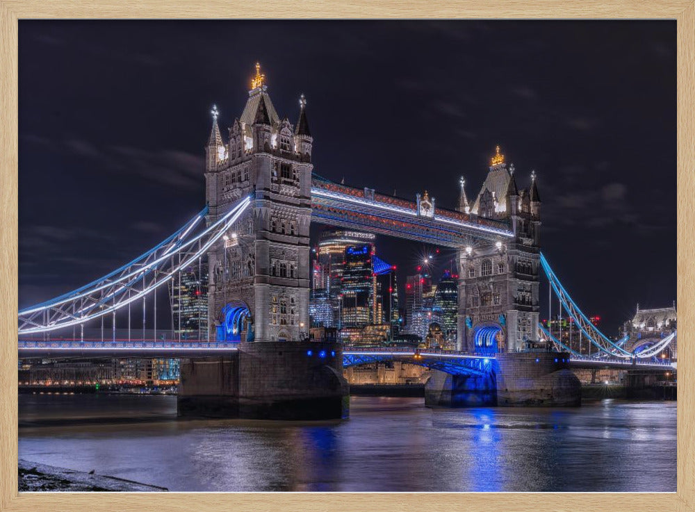 A vibrant night photograph of the iconic Tower Bridge in London, illuminated with bright white and electric blue lights. The dark River Thames in the foreground reflects the bridge and the lit-up modern city skyline in the background, all under a dark, cloudy sky. The image is enclosed in a silver frame. Decor