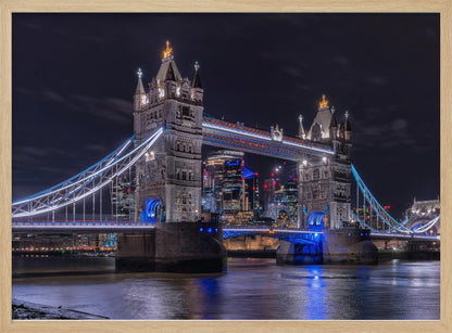 A vibrant night photograph of the iconic Tower Bridge in London, illuminated with bright white and electric blue lights. The dark River Thames in the foreground reflects the bridge and the lit-up modern city skyline in the background, all under a dark, cloudy sky. The image is enclosed in a silver frame. Decor