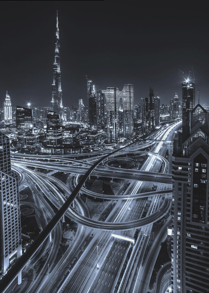 A cool-toned black and white photograph of the Dubai skyline at night. The iconic Burj Khalifa stands tall in the background, while the foreground is dominated by a massive, complex highway interchange with light trails from moving cars, creating a sense of motion and modernity. Poster