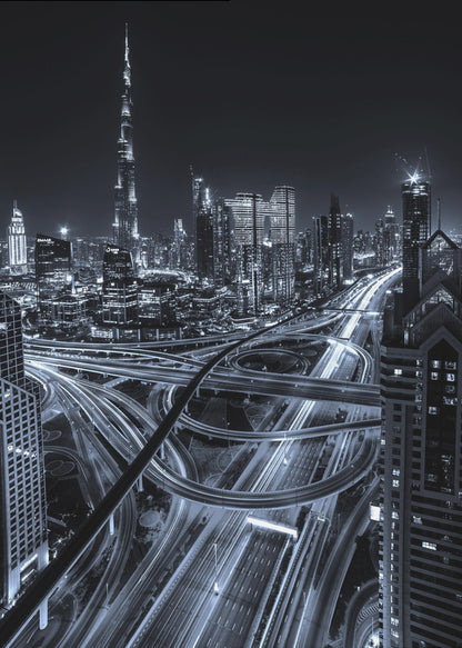 A cool-toned black and white photograph of the Dubai skyline at night. The iconic Burj Khalifa stands tall in the background, while the foreground is dominated by a massive, complex highway interchange with light trails from moving cars, creating a sense of motion and modernity. Poster