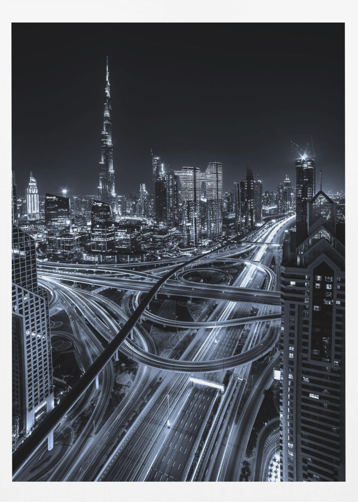 A cool-toned black and white photograph of the Dubai skyline at night. The iconic Burj Khalifa stands tall in the background, while the foreground is dominated by a massive, complex highway interchange with light trails from moving cars, creating a sense of motion and modernity. Poster