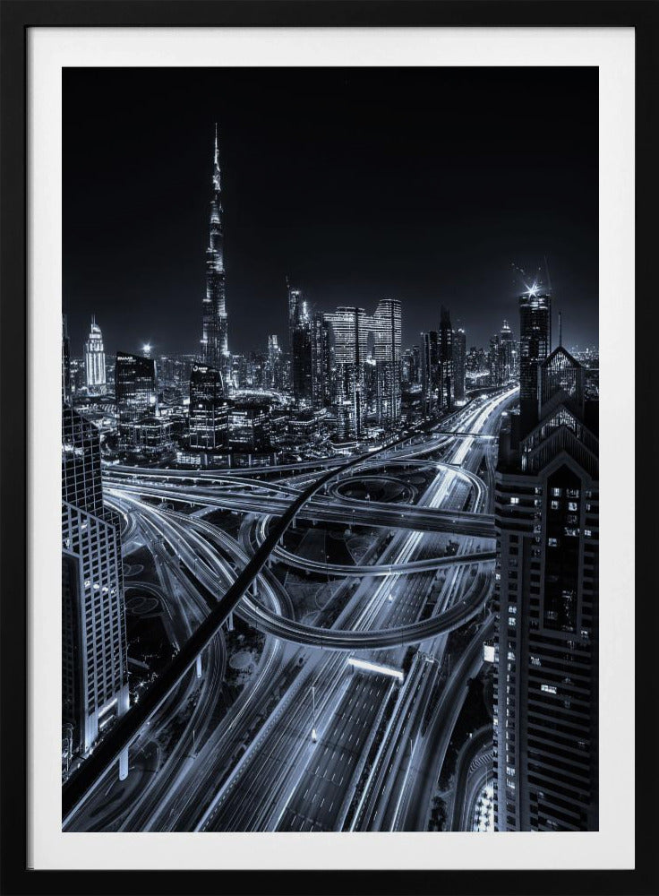 A cool-toned black and white photograph of the Dubai skyline at night. The iconic Burj Khalifa stands tall in the background, while the foreground is dominated by a massive, complex highway interchange with light trails from moving cars, creating a sense of motion and modernity. Poster